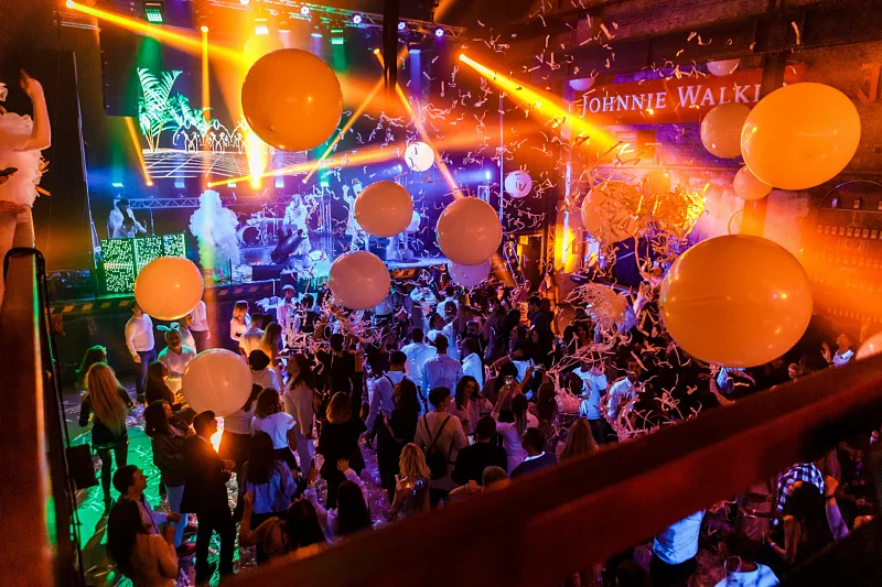 Packed nightclub dance floor with orange balloons and stage lights, seen from the balcony
